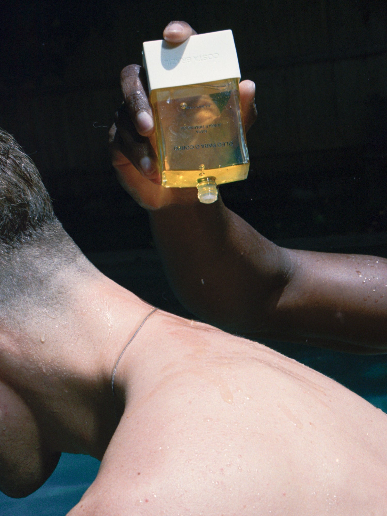 COSTA BRAZIL Kaya Jungle Firming Body Oil – lifestyle image of the body oil bottle held over wet skin by a pool