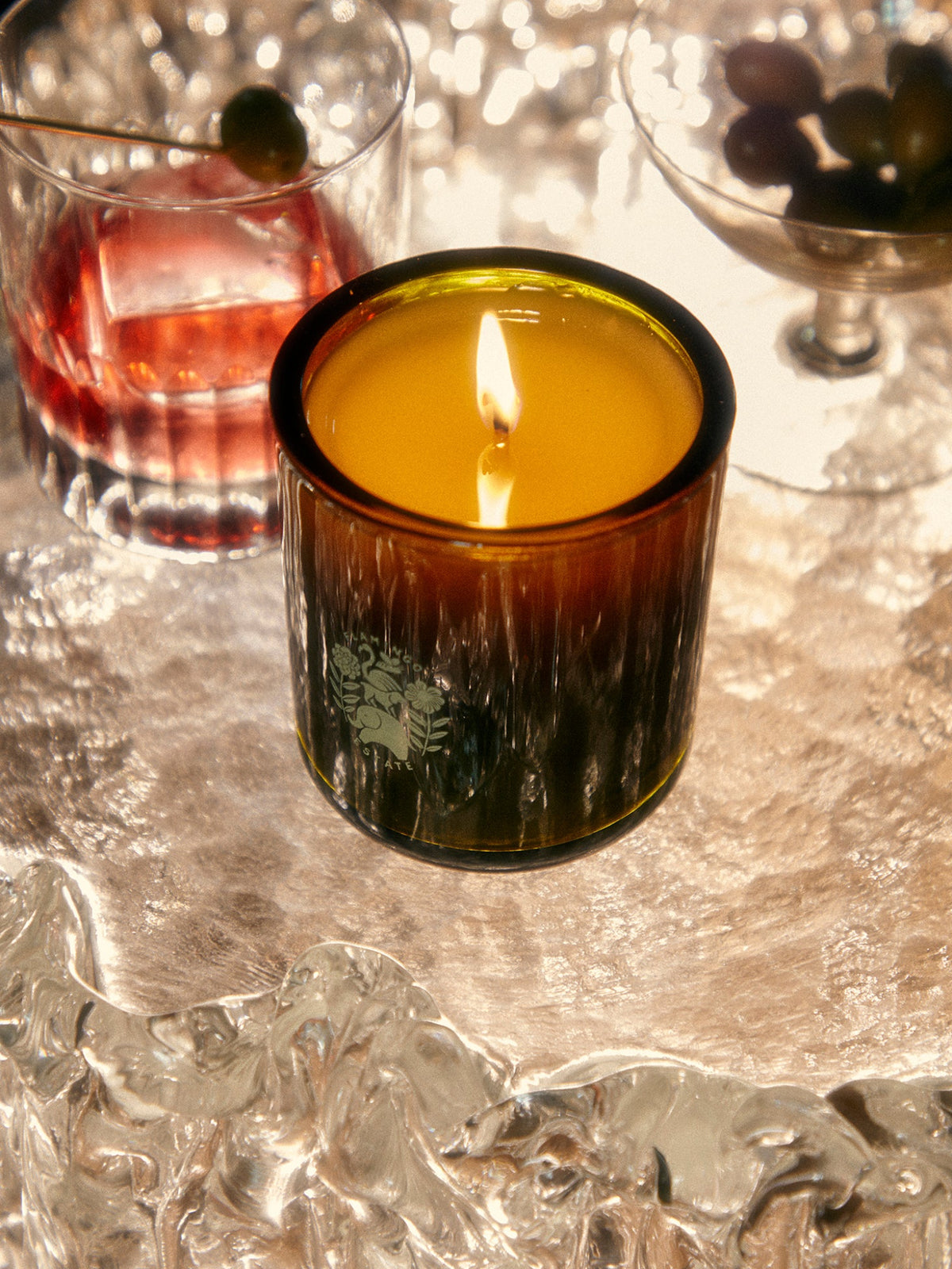 Lit Flamingo Estate Olive Tree scented candle in an amber glass vessel on a shimmering table, with drinks and olives in the background
