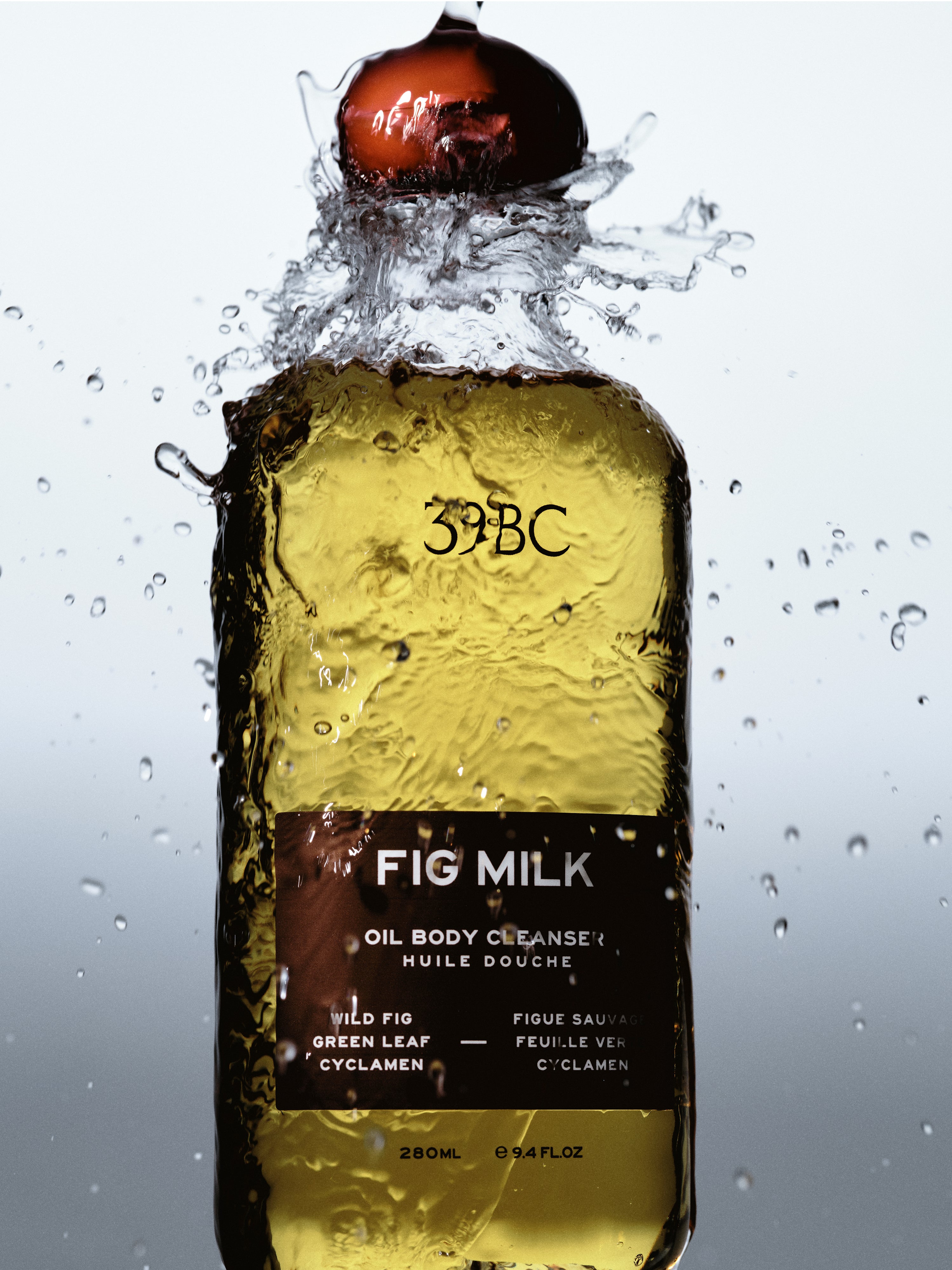 A bottle of Fig Milk oil body cleanser by 39BC, filled with golden yellow liquid and topped with a dark red fig-shaped cap, captured mid-splash with water droplets against a light gray background.
