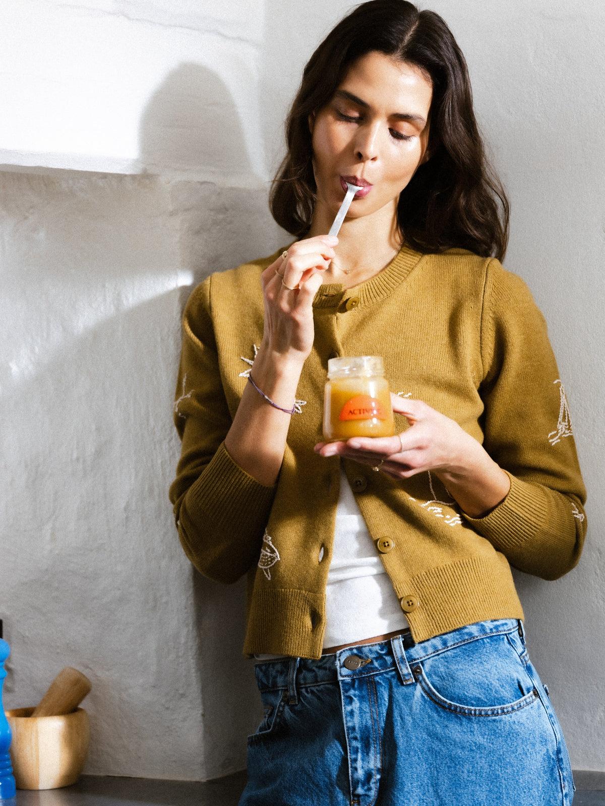 Woman tasting Active+ Manuka Honey directly from a jar with a spoon in a bright kitchen.