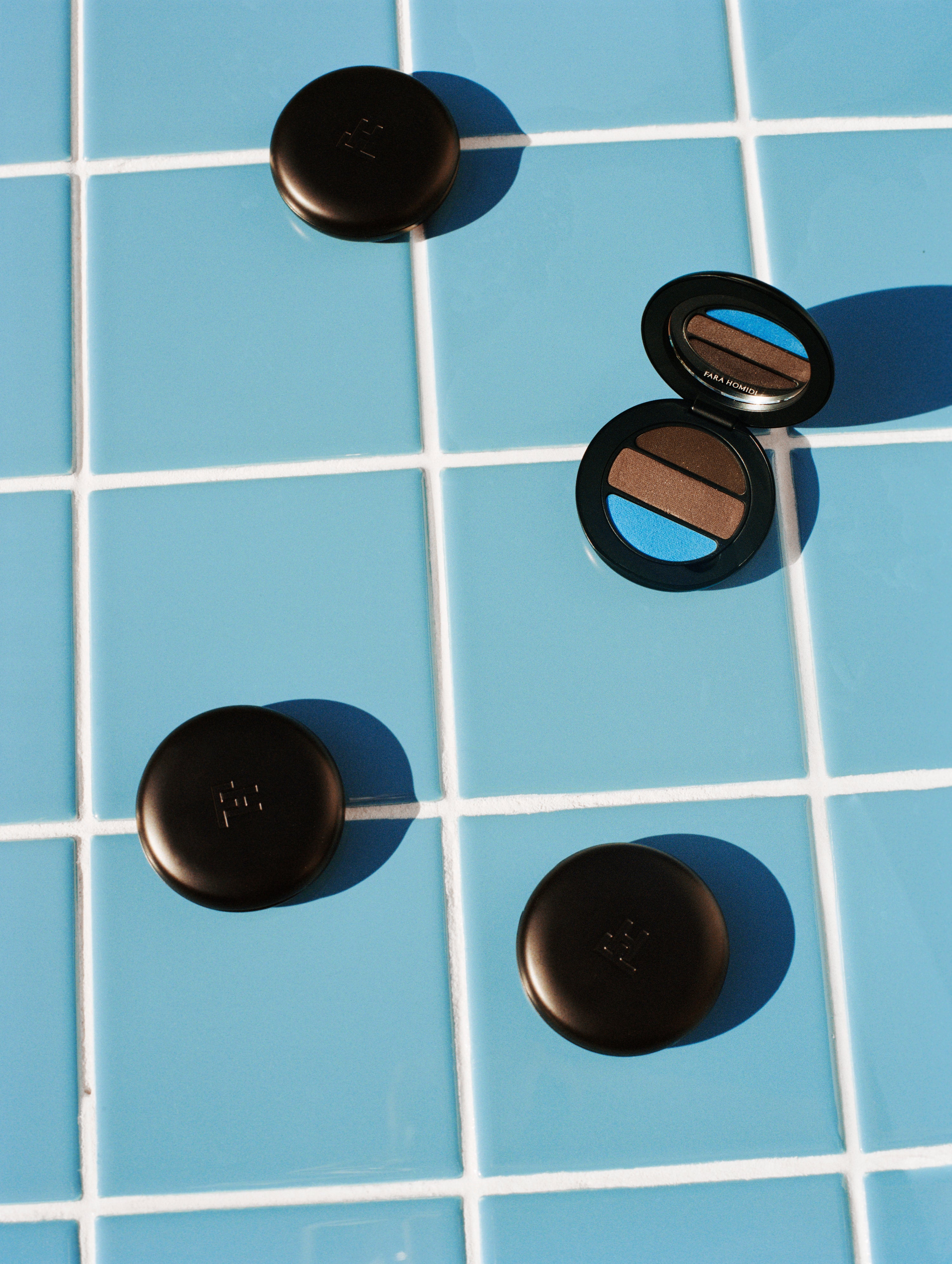 Four round black FARA HOMIDI Essential Eye Compacts, including one open eyeshadow compact showing brown and blue shades, arranged on a light blue tiled surface.