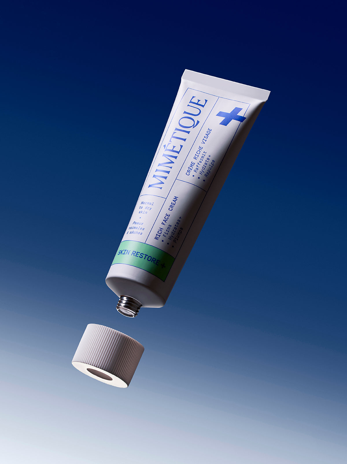 A tube of MIMÉTIQUE Skin Restore+ Rich Face Cream floating diagonally above its cap against a blue gradient background