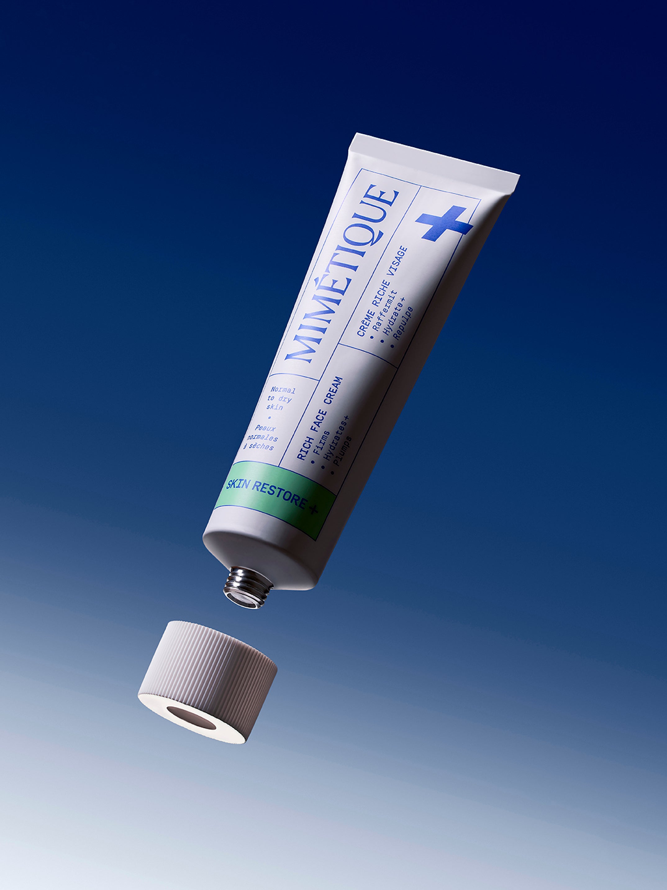 A tube of MIMÉTIQUE Skin Restore+ Rich Face Cream floating diagonally above its cap against a blue gradient background.