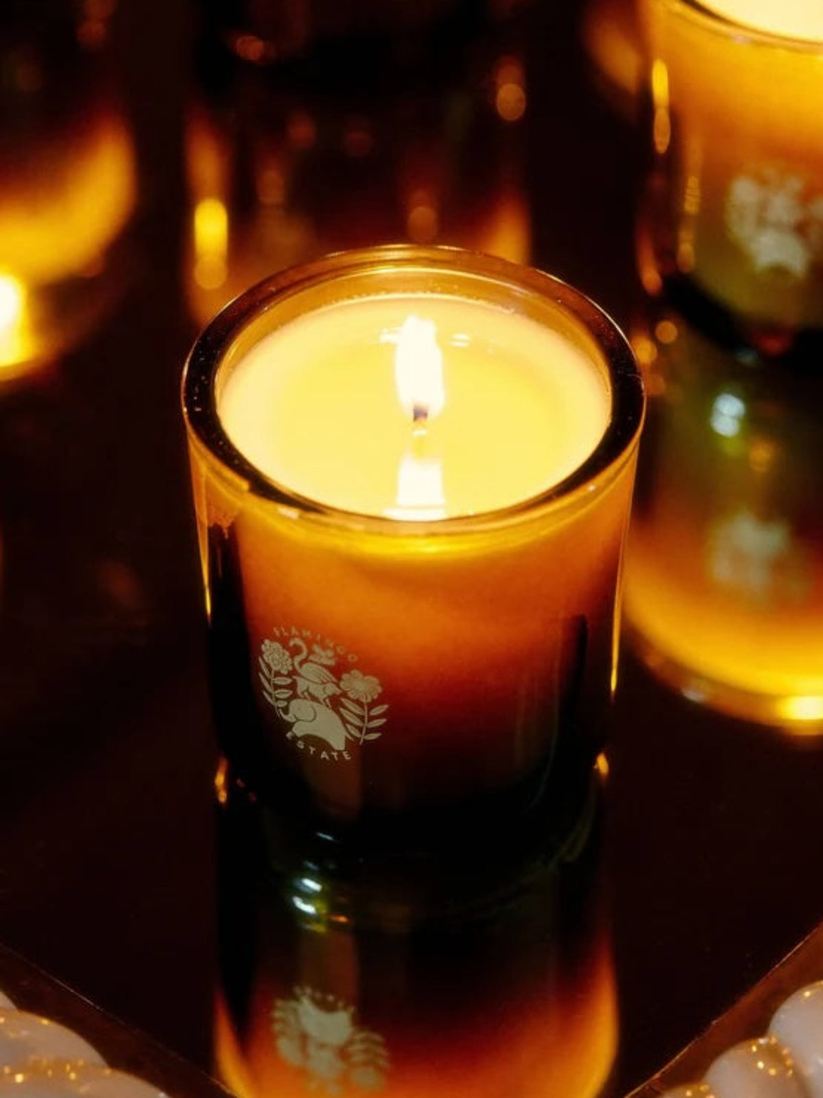 Flamingo Estate Clarity Candle in an amber glass vessel with a lit wick, photographed against a warm, glowing background