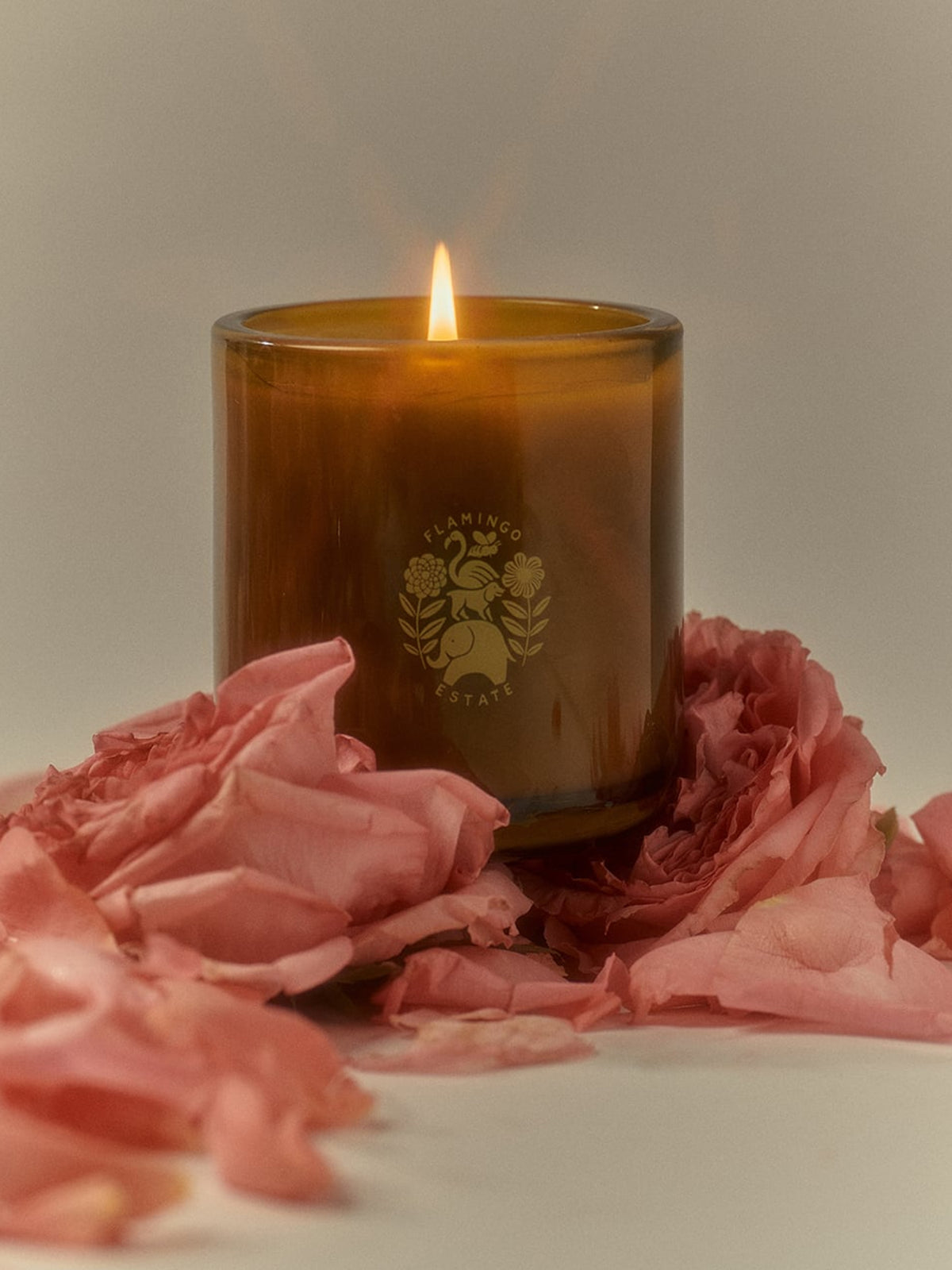 Flamingo Estate Jasmine Rose Candle in an amber glass vessel with a lit wick, surrounded by pink rose petals