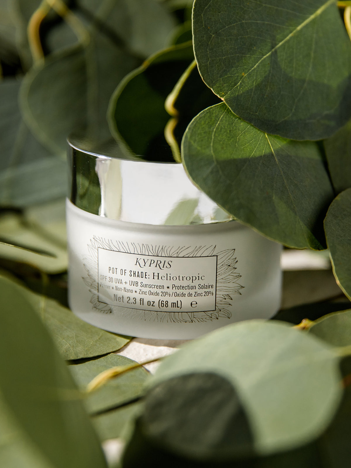 KYPRIS Pot of Shade Heliotropic SPF 30 sunscreen and primer jar surrounded by lush green leaves.
