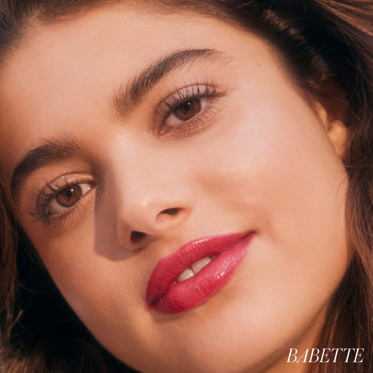 Model wearing RMS Beauty LipLights Cream Lip Gloss in Babette, showcasing glossy coral-red lips