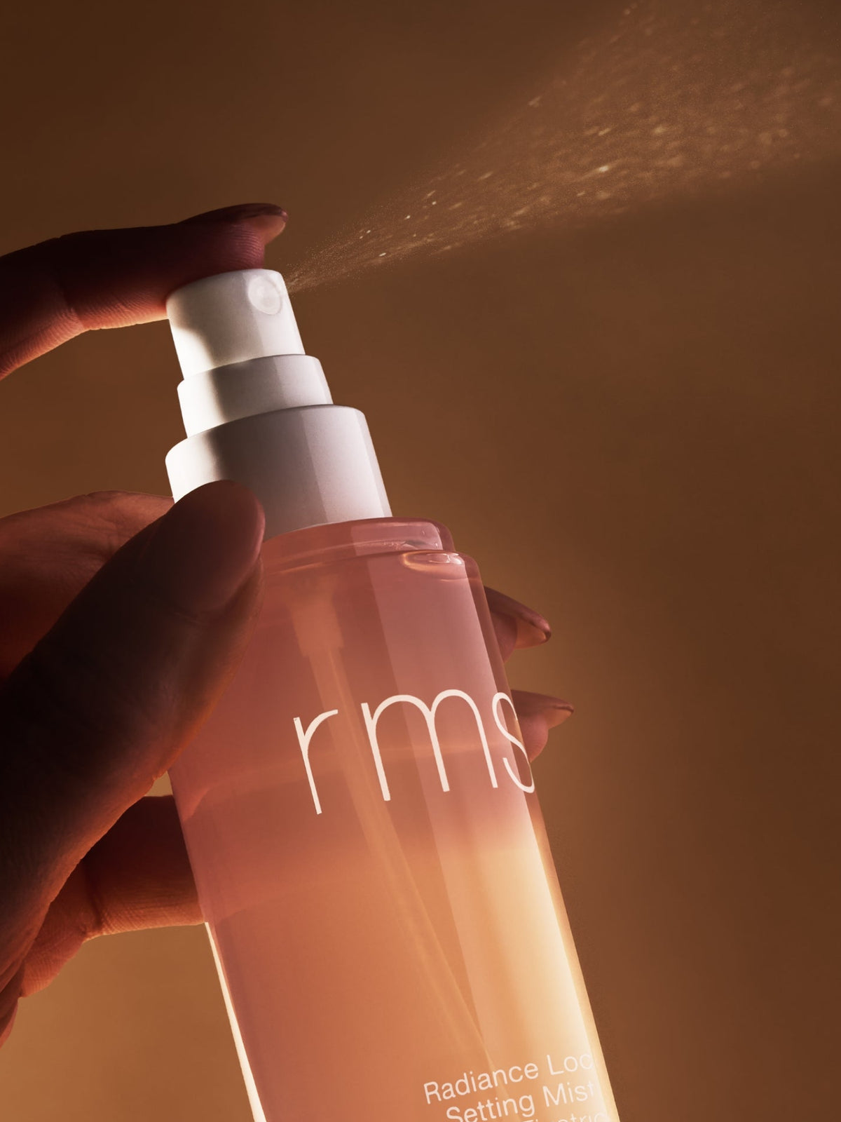 Close-up of a hand holding RMS Beauty Radiance Lock Setting Mist as warm light illuminates the peach-toned bottle
