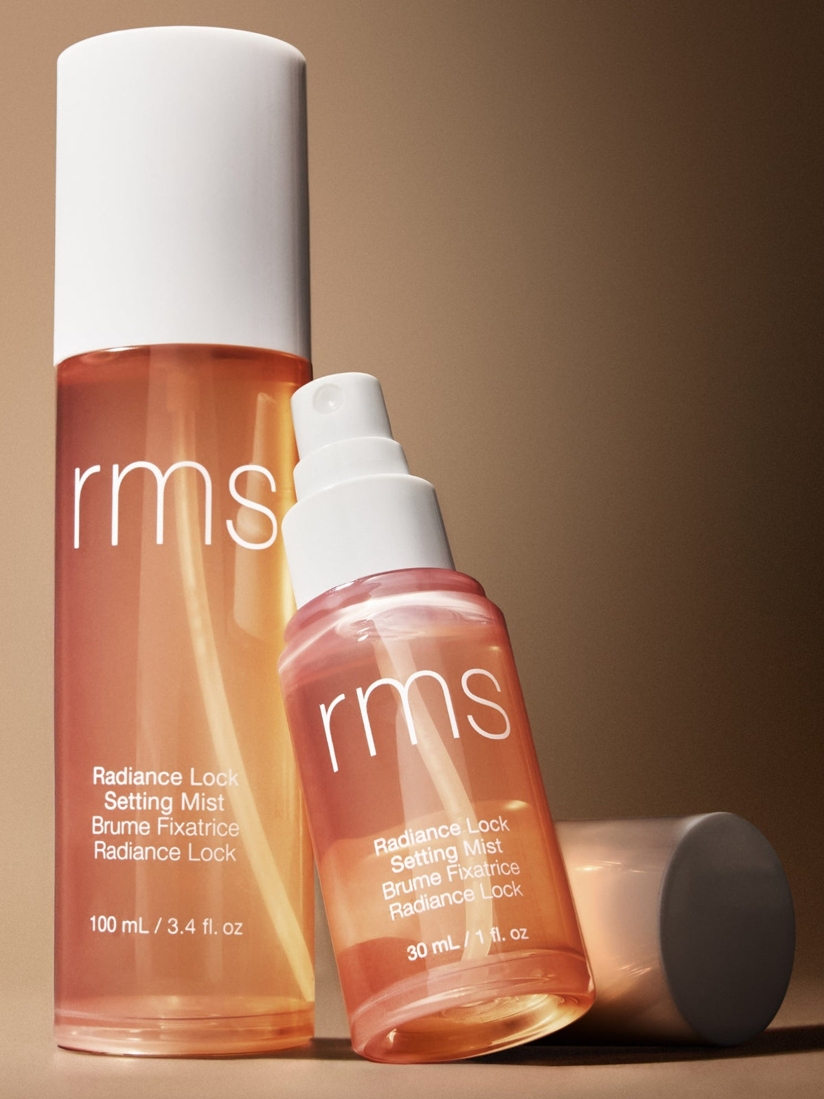 RMS Beauty Radiance Lock Setting Mist 100 ml and 30 ml spray bottles displayed together against a warm brown background