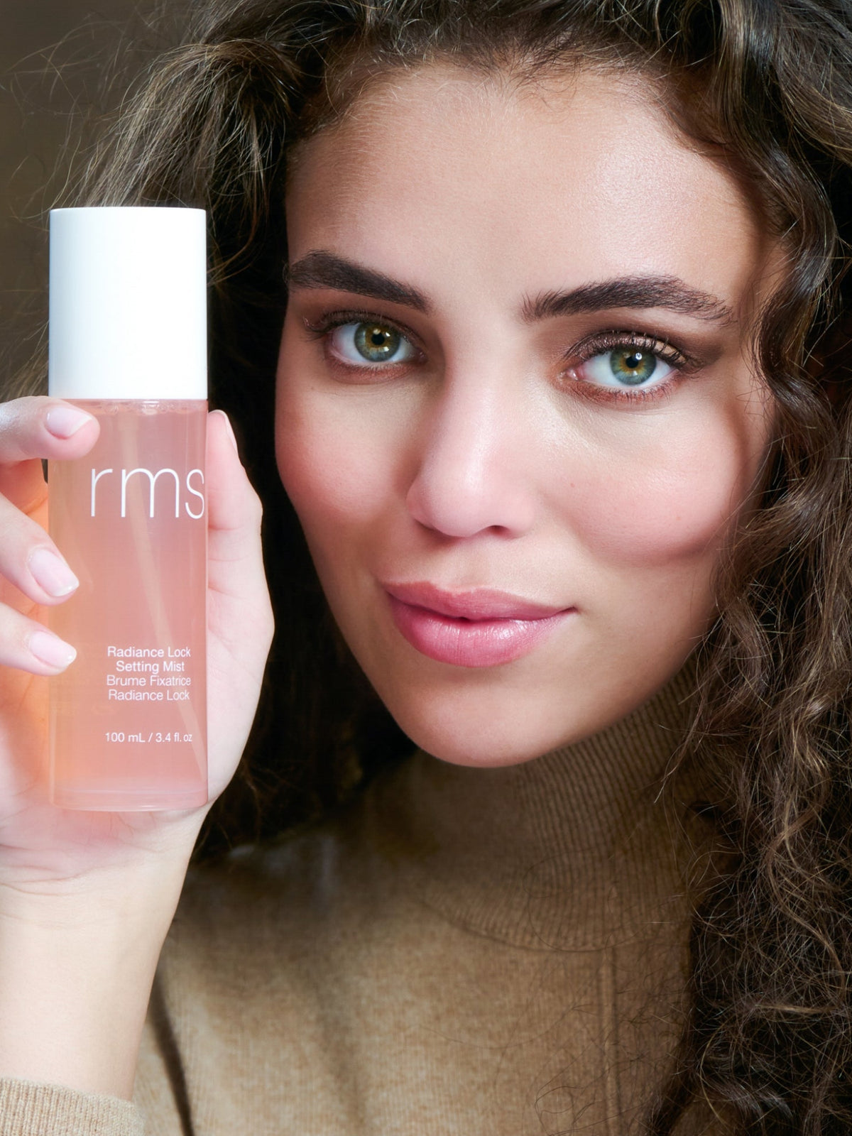 Model holding RMS Beauty Radiance Lock Setting Mist 100 ml bottle beside her face