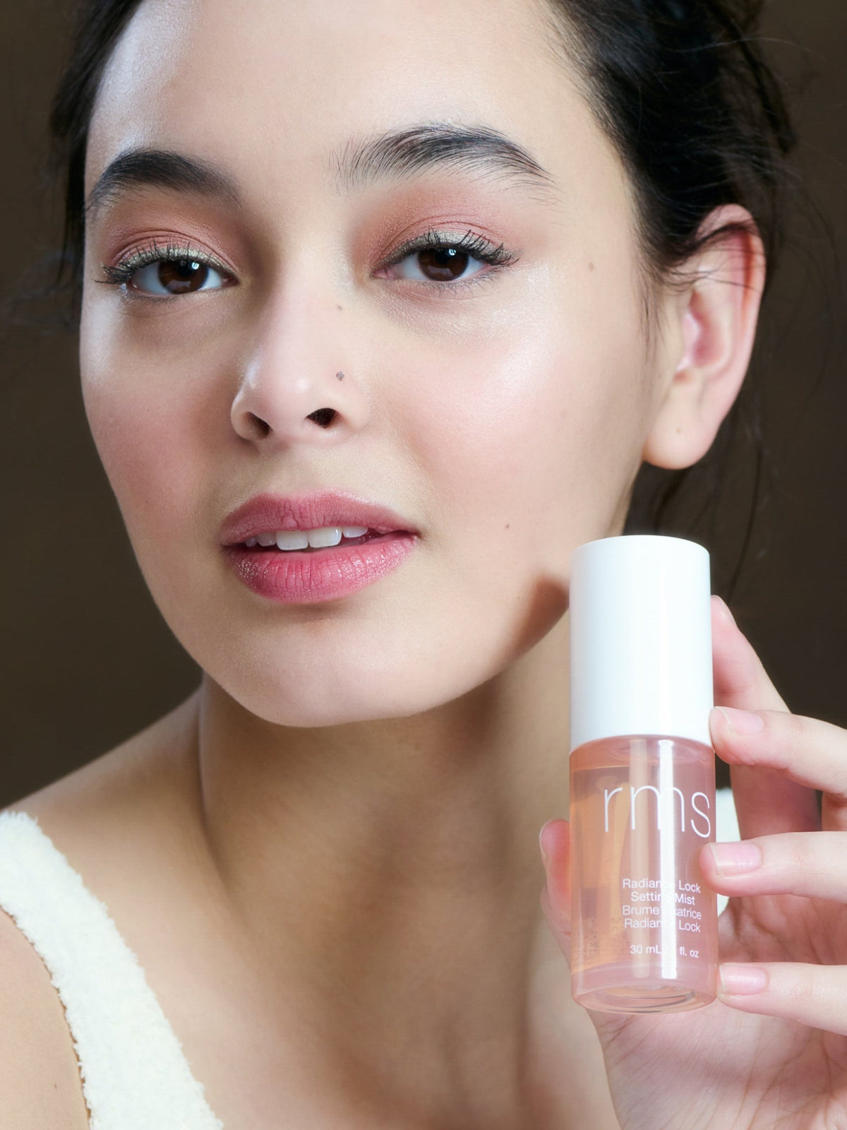Model holding RMS Beauty Radiance Lock Setting Mist 100 ml bottle and showcasing her radiant complexion