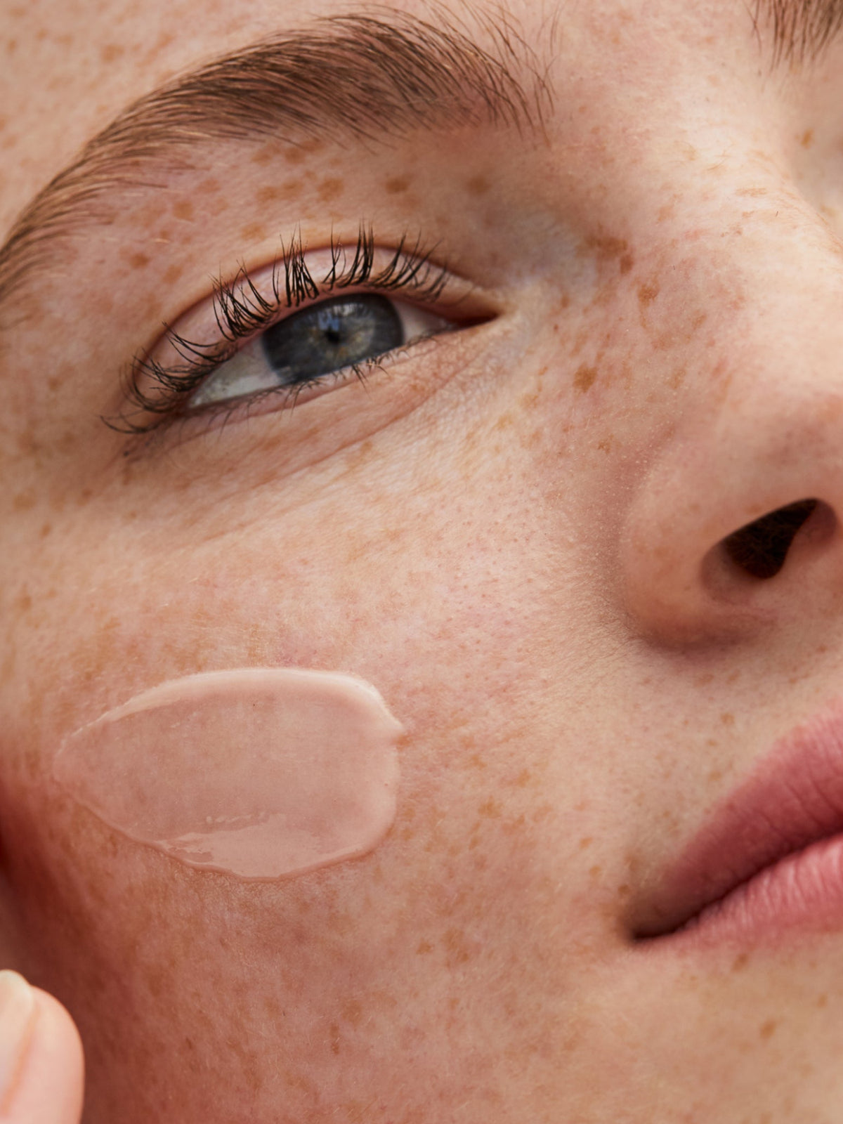 Close-up of a woman applying RMS Beauty ReEvolve Radiance Locking Primer to her cheek, showing the lightweight gel texture on the skin